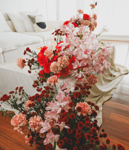Cascading floral arrangement with red roses, pink carnations, alstroemeria, and burgundy chrysanthemums on a draped table in a cozy living room setting.