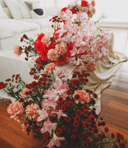 A lavish floral arrangement featuring red roses, pink alstroemeria, peach carnations, and red spray chrysanthemums, placed on a draped table in a modern living room.