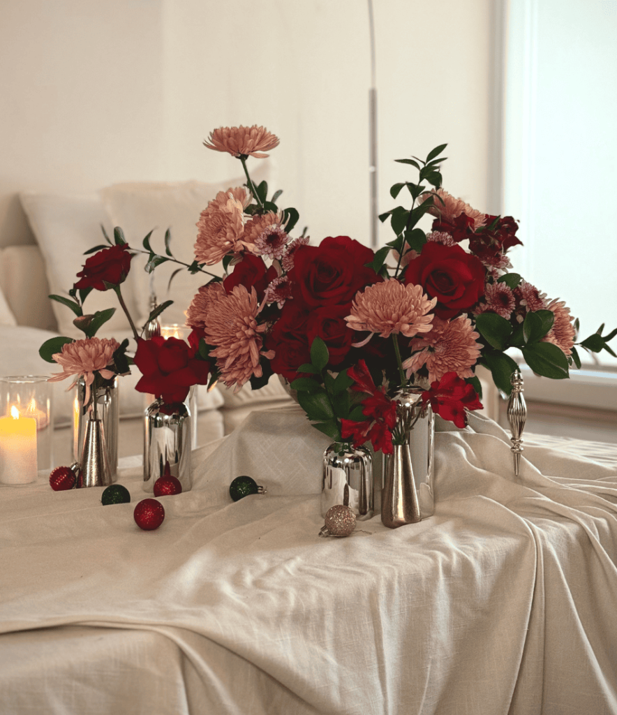 A holiday floral arrangement featuring lush red roses, peach chrysanthemums, green foliage, and playful ornaments hung on floral stems, set on a draped white fabric with festive accents.