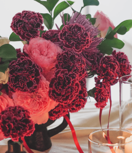 Close-up of a gothic floral arrangement with pink and dark purple carnations, cream spray roses, purple spider flowers, and ruscus in a black vase, accented with red bows.