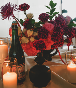 Gothic floral tablescape featuring pink and dark purple carnations, cream spray roses, a green wine bottle, candles, and a black vase.