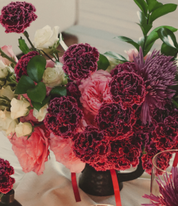 Overhead view of a gothic floral arrangement with pink and dark purple carnations, cream spray roses, ruscus, and purple spider flowers in a black vase.