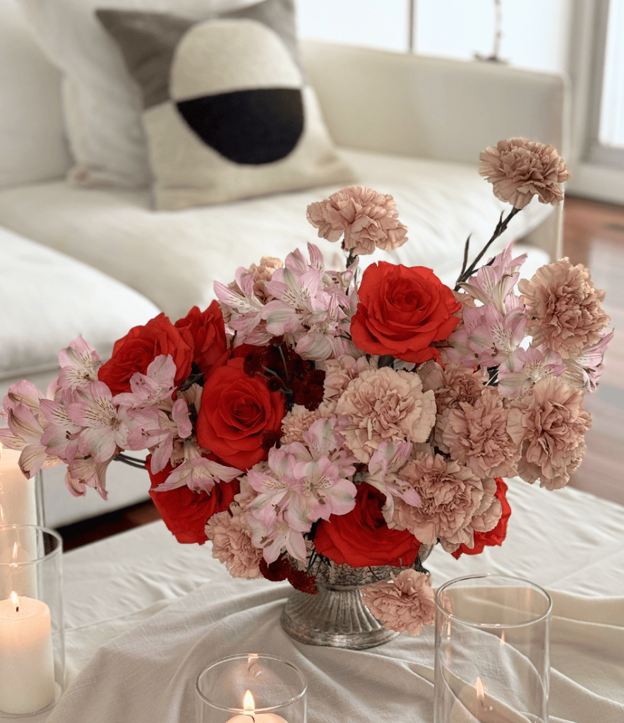 Floral arrangement with red roses, pink carnations, and alstroemeria in a silver vase, surrounded by glass candle holders.