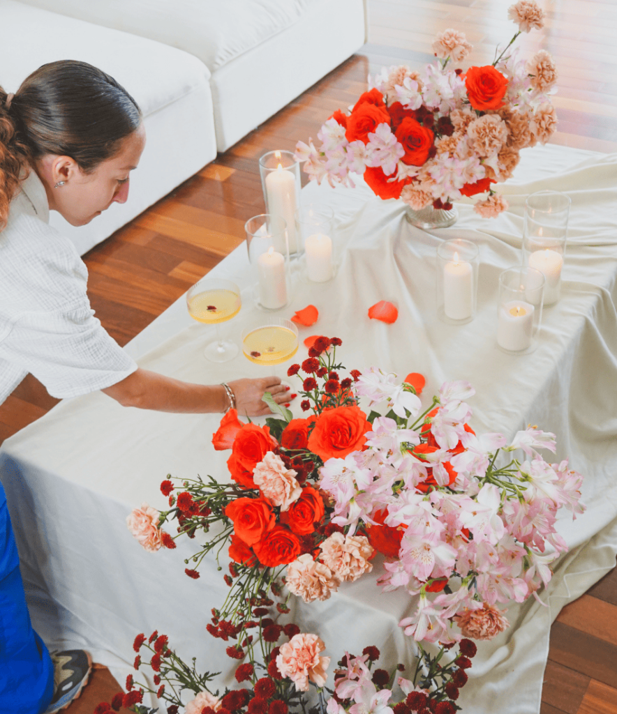 A florist arranging a floral display with red roses, pink carnations, and alstroemeria on a table with candles and champagne glasses.