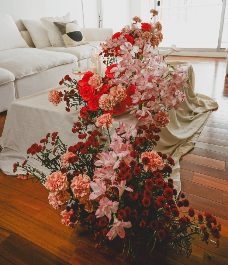 Cascading floral arrangement with red roses, pink carnations, alstroemeria, and burgundy chrysanthemums on a draped table in a cozy living room setting.