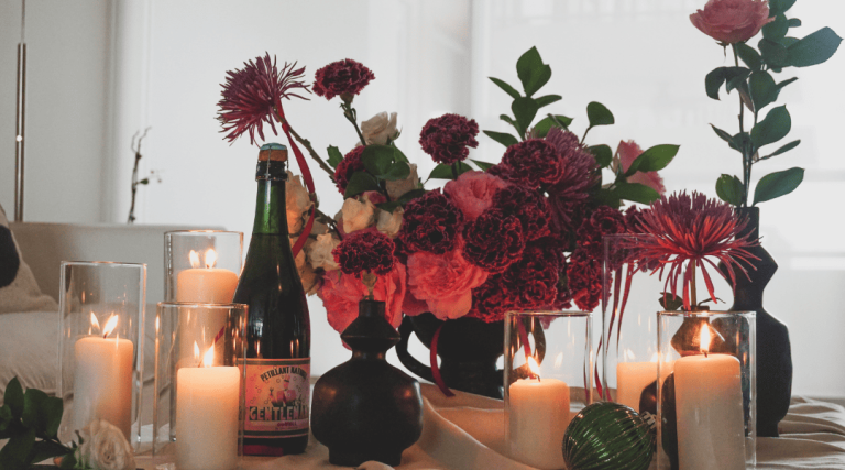 A Gothic Christmas floral arrangement featuring deep red carnations, pink roses, white spray roses, and purple spider mums in black vases, accented with green ruscus, glowing candles, and a sparkling holiday ornament.