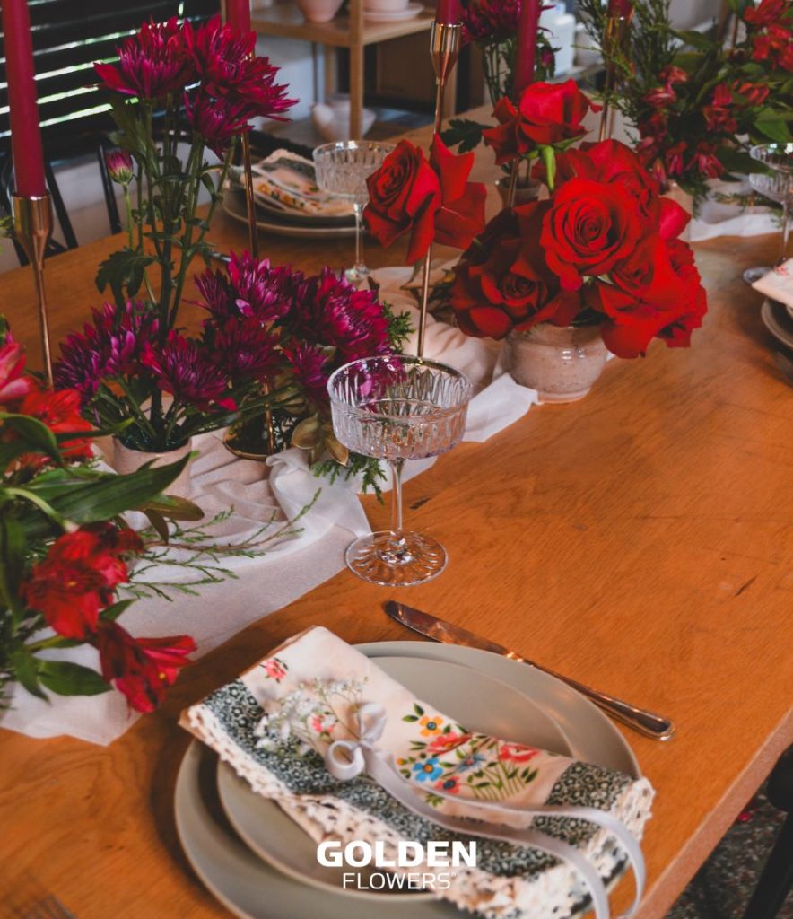 Holiday table arranged with red roses, purple chrysanthemums, alstroemeria, and greenery beside glassware and red candles.